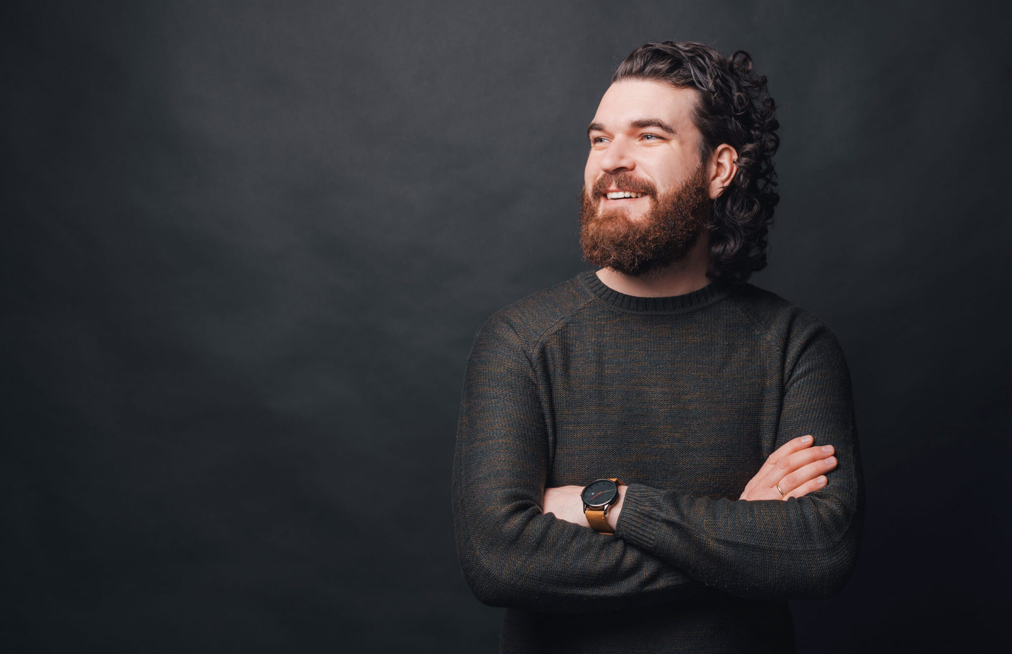 Handsome man with beard and long curly hair standing over dark background with crossed arms and looking away