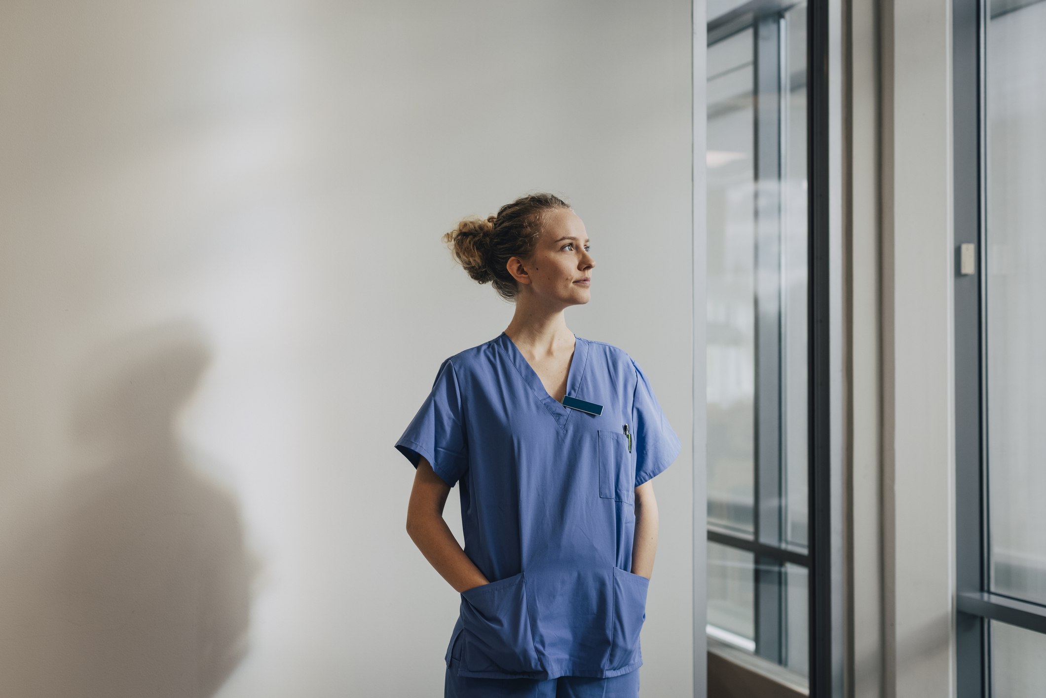 Thoughtful young nurse standing with hands in pockets against wall at hospital